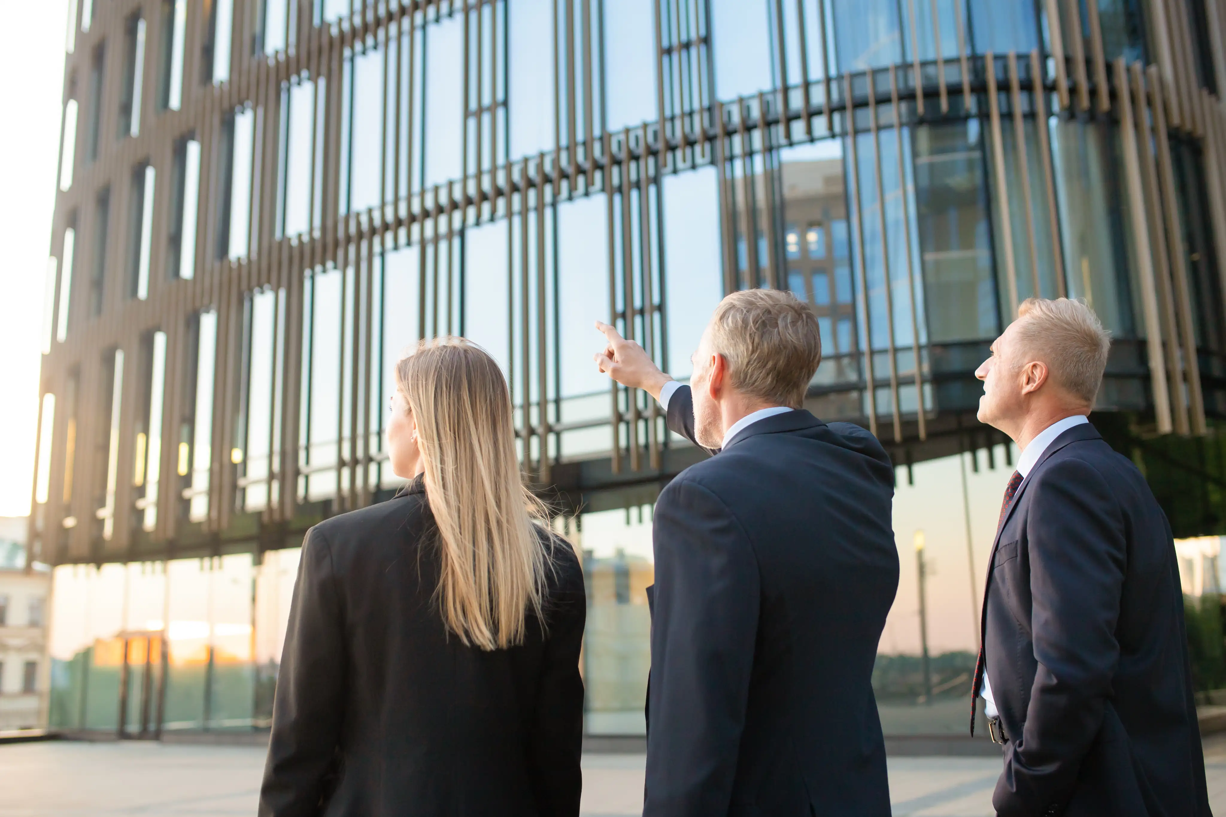 Three business professionals in suits standing outside a modern office building, with one person pointing upward, symbolizing leadership and vision