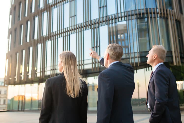 Three business professionals in suits standing outside a modern office building, with one person pointing upward, symbolizing leadership and vision