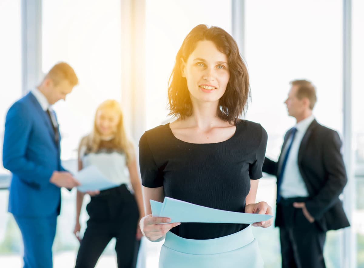 Business professional holding documents and smiling in focus, with three colleagues conversing in the background inside a modern office