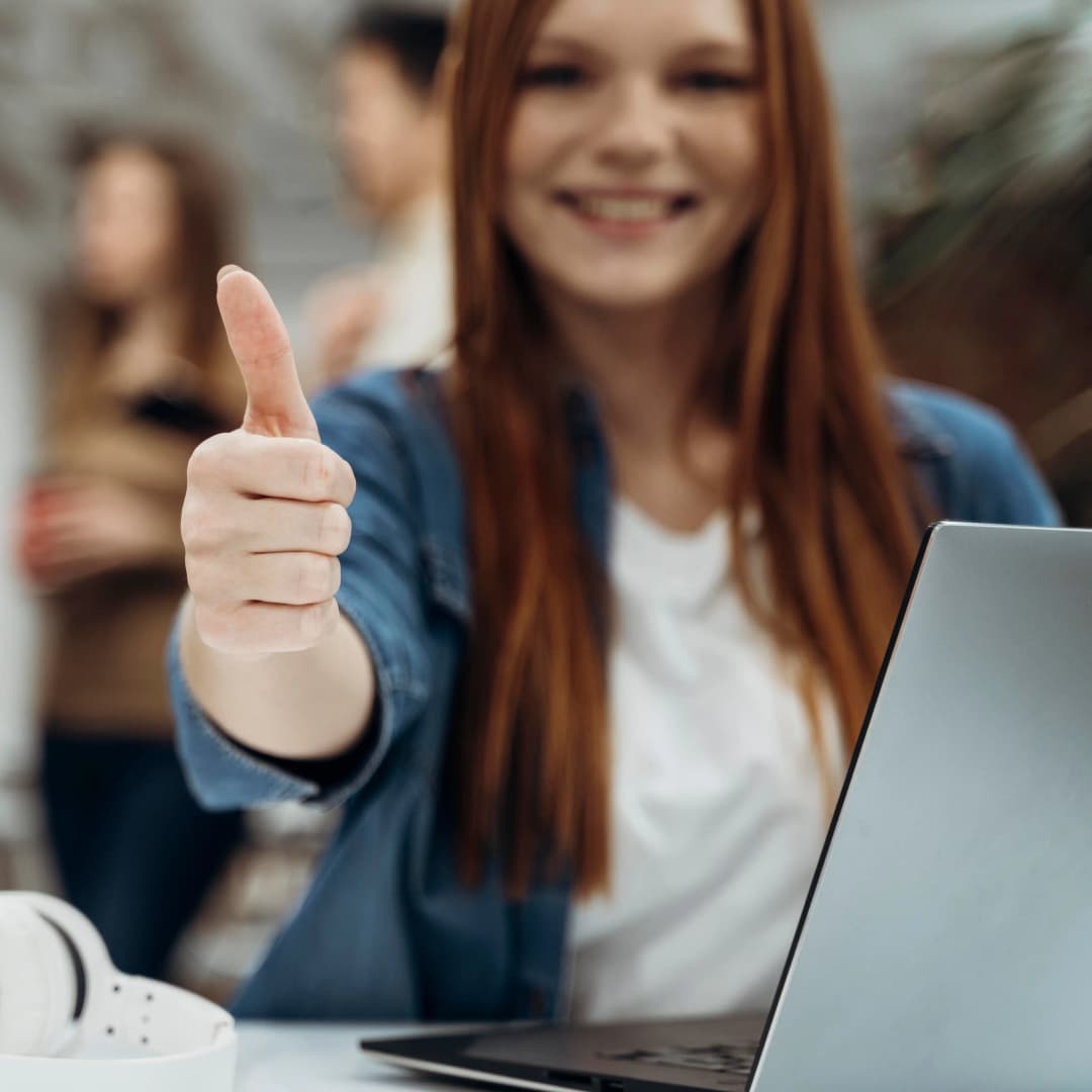 Three business professionals in an office setting, one in focus smiling and giving a thumbs up at a desk with a laptop and wireless headphones, illustrating confidence and satisfaction in choosing the best business structure.