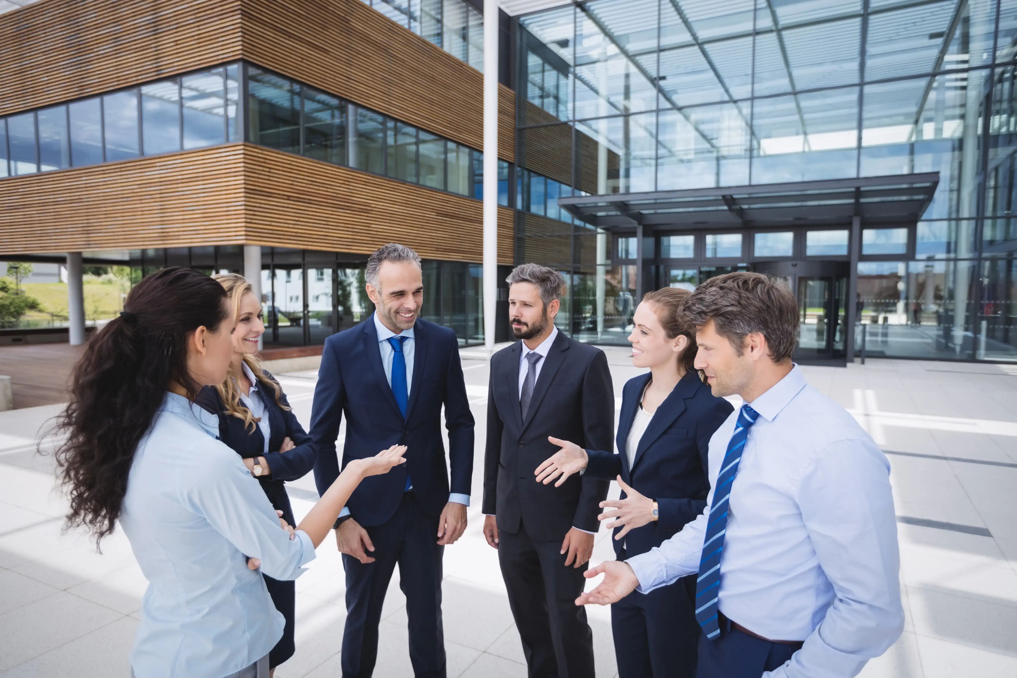 Group of six business professionals in formal attire having a discussion and smiling outside a modern office building